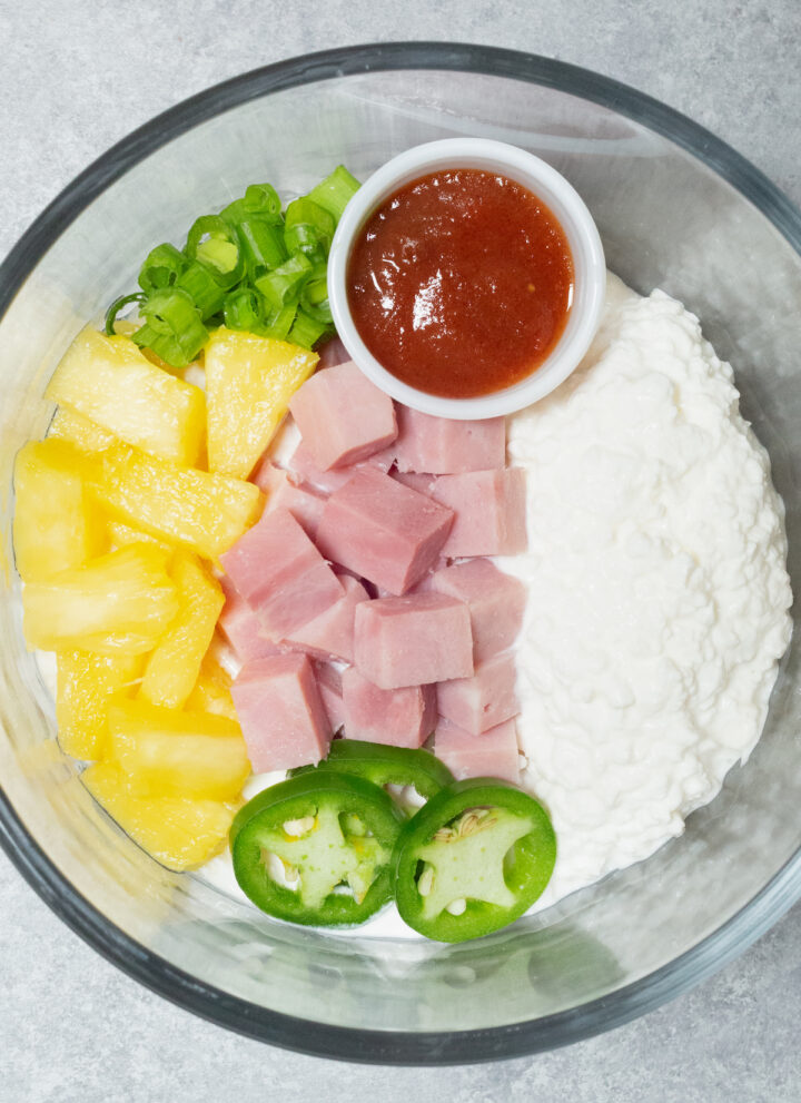 A clear glass bowl is filled with cottage cheese, ham chunks, pineapple chunks, sliced green onions, and sliced jalapenos.