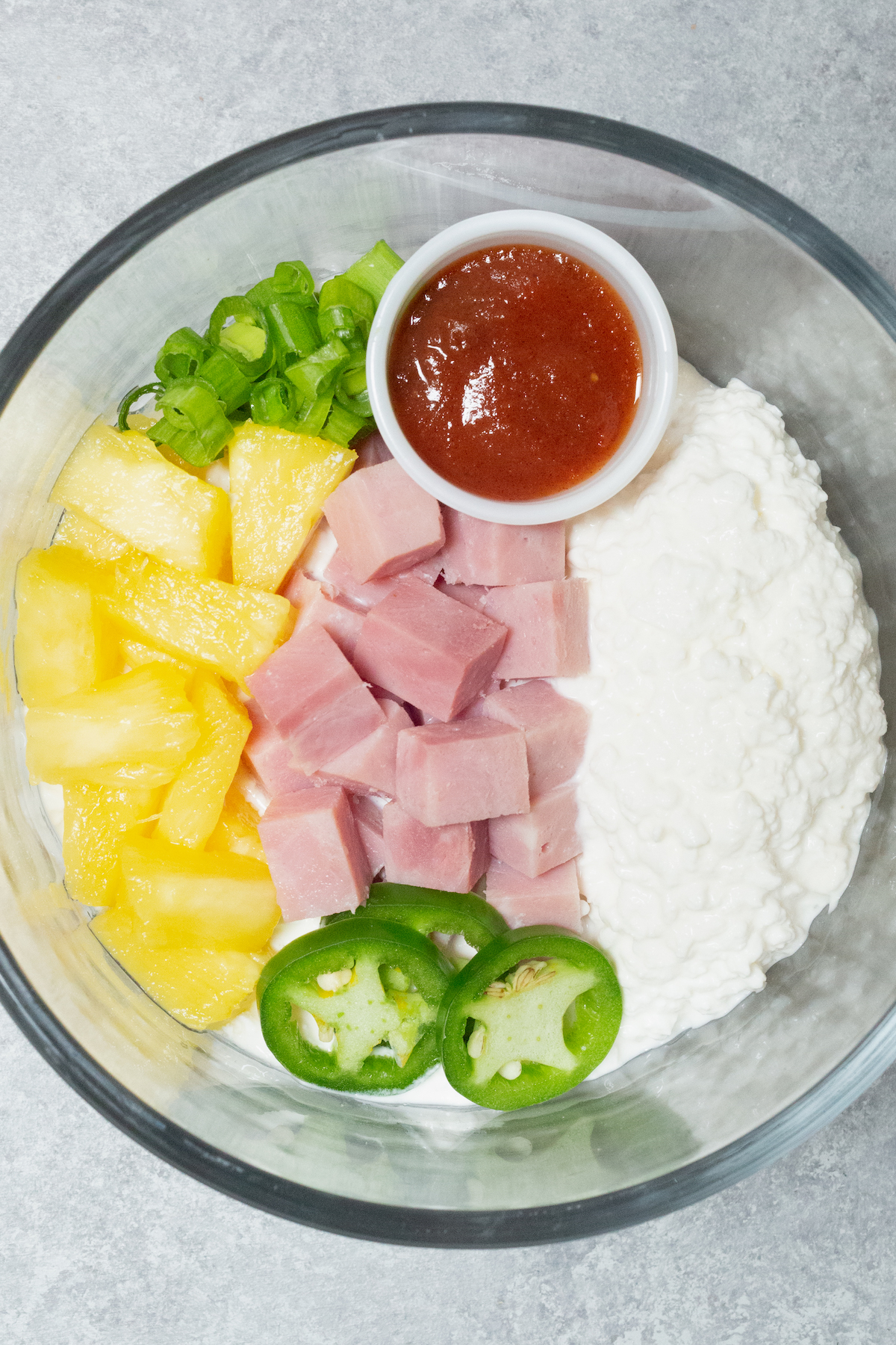A clear glass bowl is filled with cottage cheese, ham chunks, pineapple chunks, sliced green onions, and sliced jalapenos.