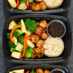 Overhead view of three black plastic meal prep containers each filled with brown rice, browned teriyaki tofu cubes, and steamed vegetables, along with a 2 oz container of teriyaki sauce.