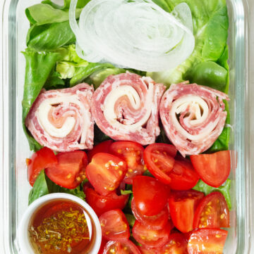 Lunch meat and cheese are rolled in to pinweheels and placed on top of a bed of greens next to sliced cherry tomatoes and sliced white onion. There is a 1 ounce container with oil and vinegar in the left bottom corner of the meal prep container.