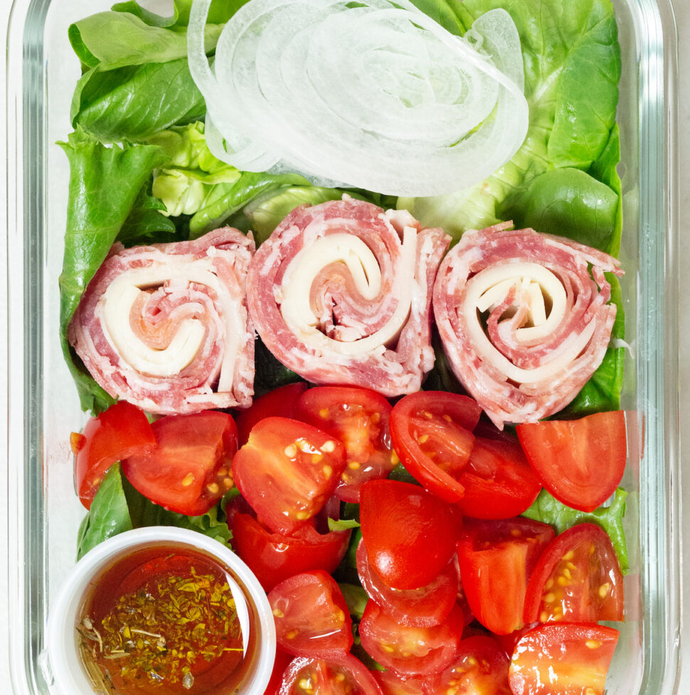 Lunch meat and cheese are rolled in to pinweheels and placed on top of a bed of greens next to sliced cherry tomatoes and sliced white onion. There is a 1 ounce container with oil and vinegar in the left bottom corner of the meal prep container.