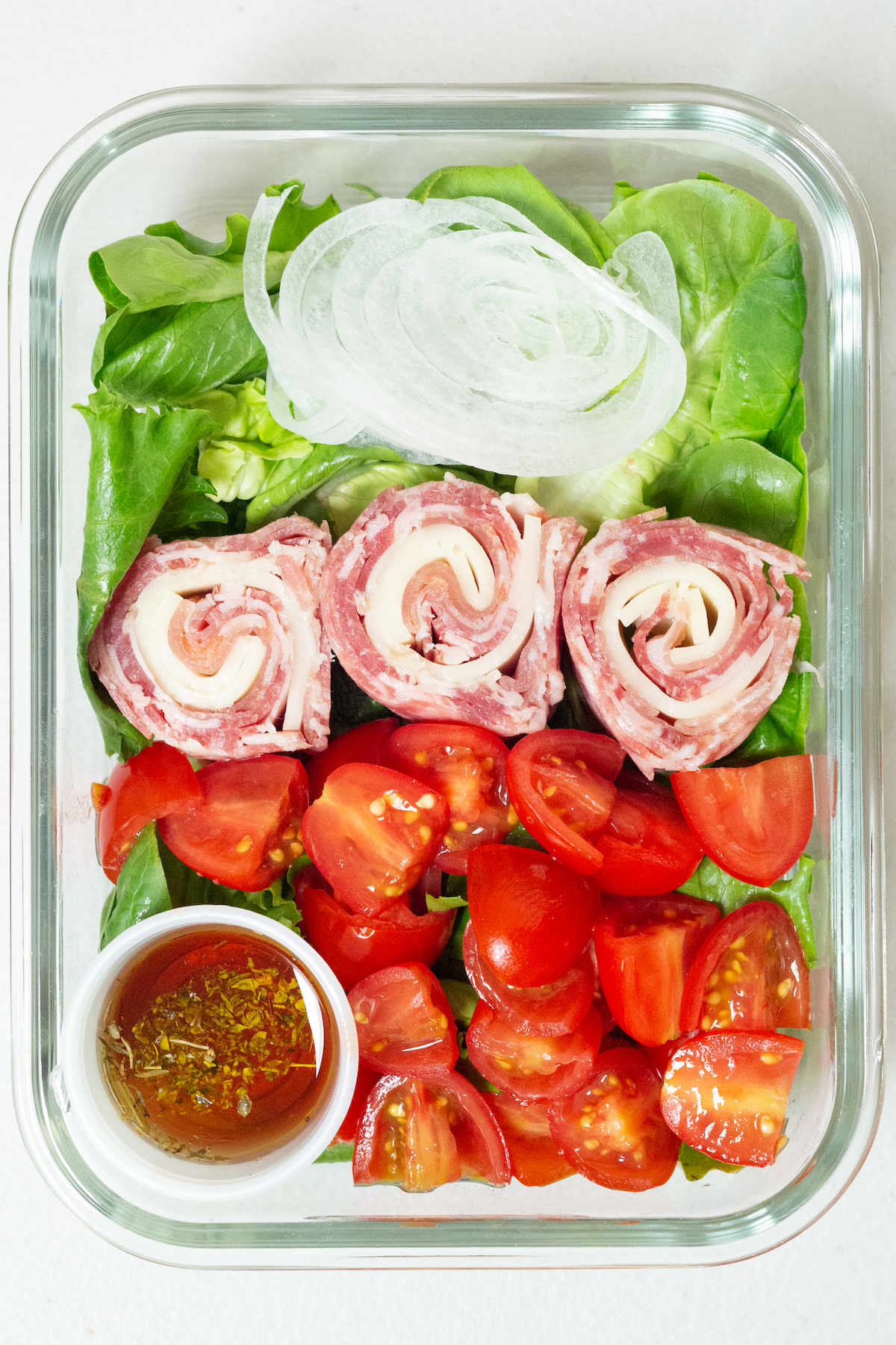 Lunch meat and cheese are rolled in to pinwheels and placed on top of a bed of greens next to sliced cherry tomatoes and sliced white onion. There is a 1 ounce container with oil and vinegar in the left bottom corner of the meal prep container.